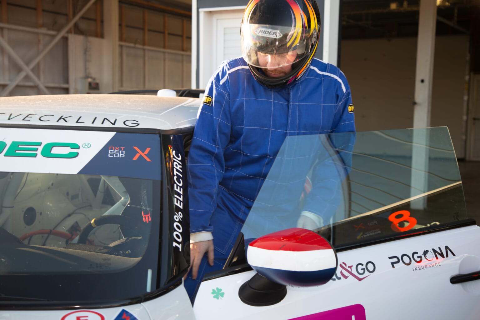 En person i blå racingdräkt och hjälm kliver in i en vit bil märkt 100% ELECTRIC med öppen dörr. Bilen har sponsorlogotyper och numret 8 på rutan.