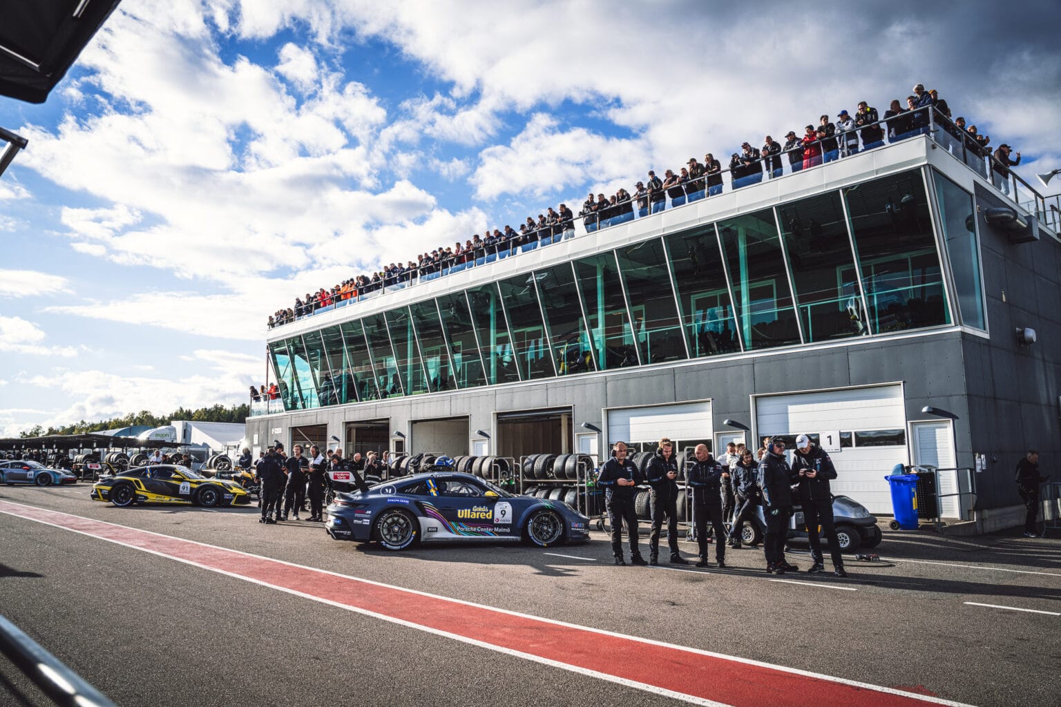 Racerbilar och förare står uppradade på en bana nära en modern glasbyggnad med stora fönster. Åskådare tittar på från taket och himlen är delvis molnig. Tävlingsdäck och garageportar syns i bakgrunden.