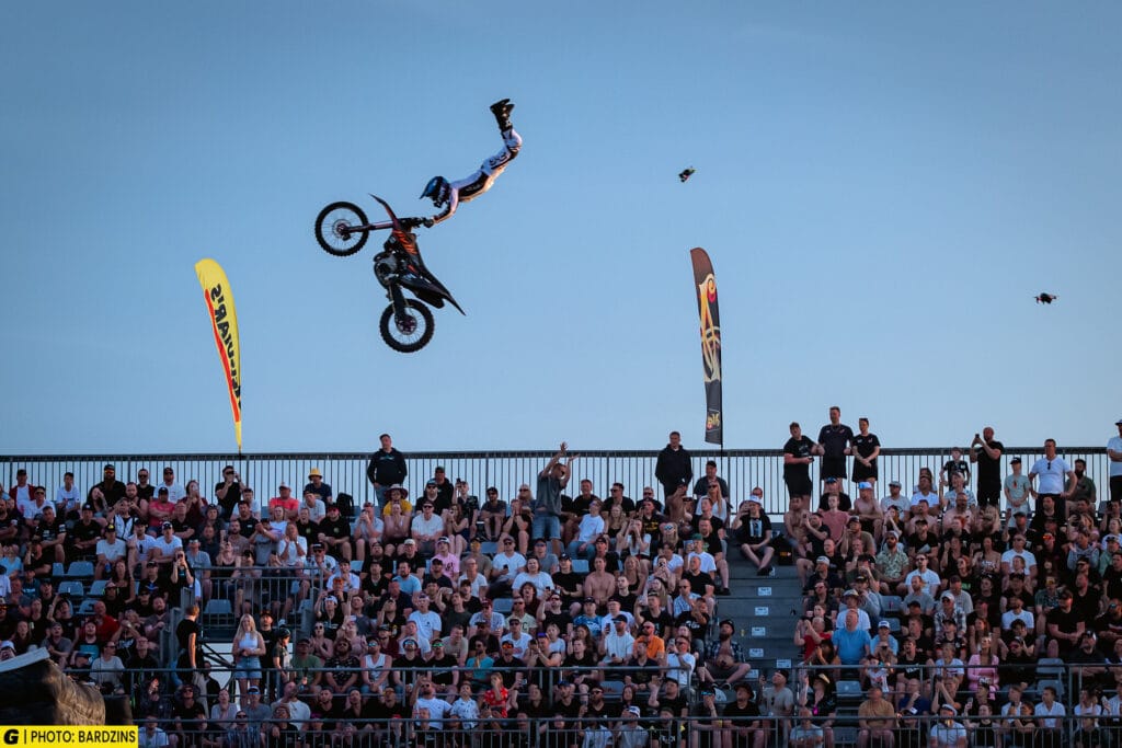 En motocrossförare utför ett stunt i luften och släpper taget om sin motorcykel ovanför en stor publik på en utomhusarena, med banderoller och en klarblå himmel i bakgrunden.