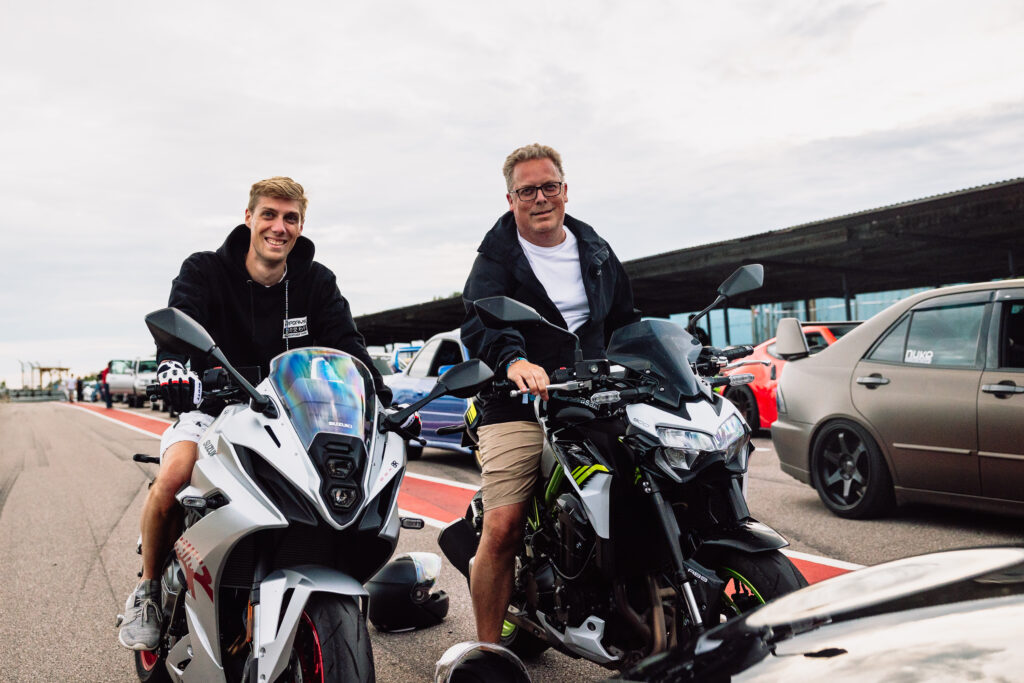 Två män sitter på motorcyklar sida vid sida på en racerbana och ler mot kameran. Bilar och andra motorcyklar står uppradade i bakgrunden under en molnig himmel.