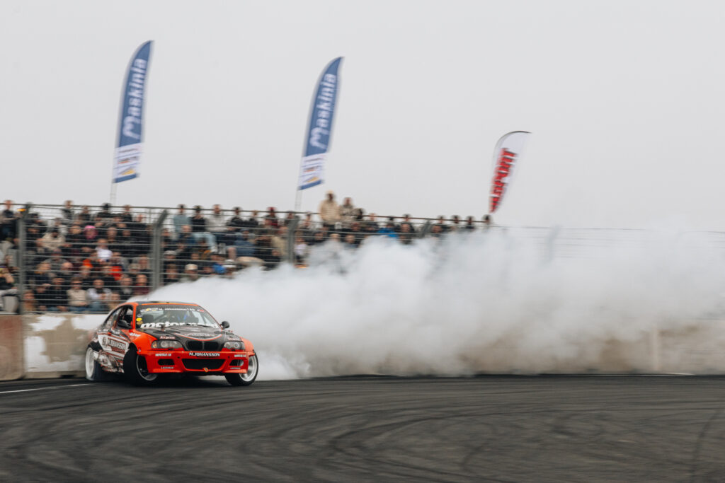 En röd racerbil driver runt ett hörn på en racerbana och skapar ett stort moln av vit rök, med en mängd åskådare och flera banderoller i bakgrunden.