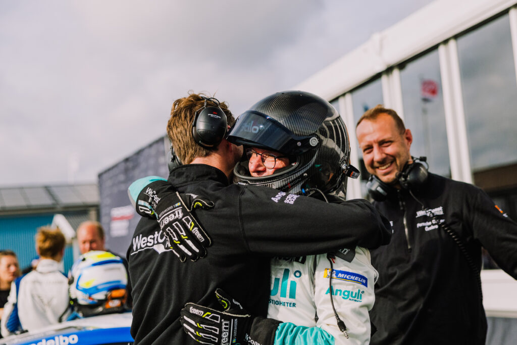 En racerförare i hjälm och tävlingsdräkt kramar om en teammedlem, båda ler glatt. En annan teammedlem står i närheten, också leende, med en racerbil och en bana i bakgrunden.