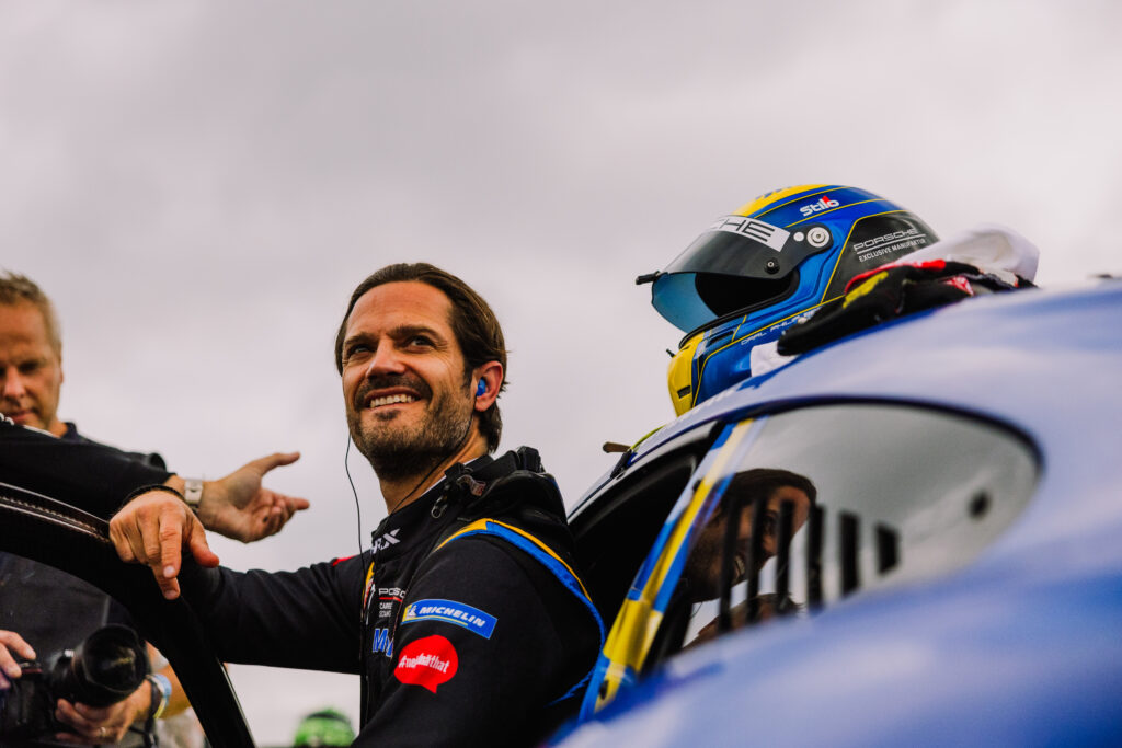 En leende racerförare står bredvid en blå bil med en racinghjälm vilande på taket, medan en annan person gestikulerar i närheten under en molnig himmel.