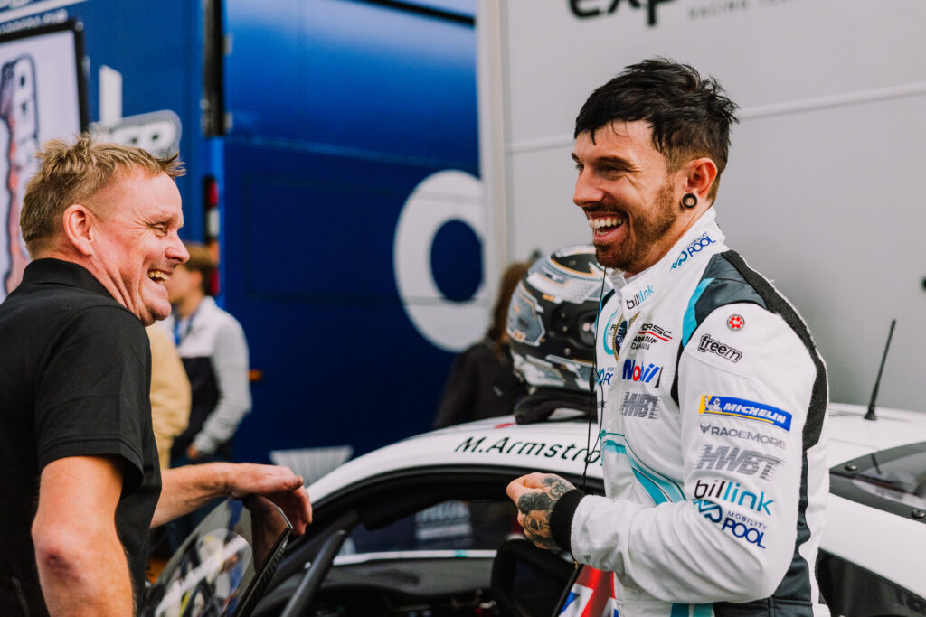 Två män står leende bredvid en racerbil; den ene bär en racingdräkt med olika sponsorlogotyper, medan den andre är vardagligt klädd. Stämningen verkar vänlig och uppsluppen, och en hjälm vilar på bilen i närheten.