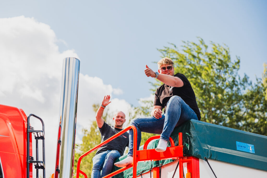 Två män sitter på en färgglad lastbil; den ena ger tummen upp och den andra vinkar, båda ler. I bakgrunden syns träd och en blå himmel med moln.