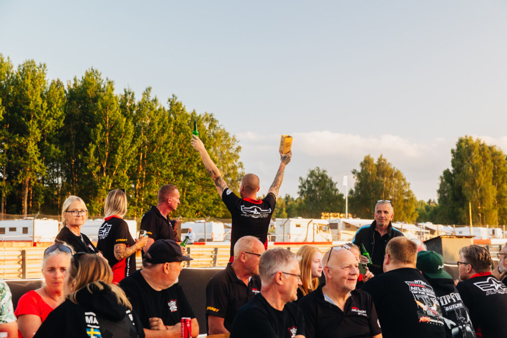 En grupp människor sitter och står utomhus nära husvagnar och träd. En person i mitten står med armarna i vädret och håller i en drink och ett mellanmål, medan de andra tittar på eller pratar i solnedgångens gyllene ljus.