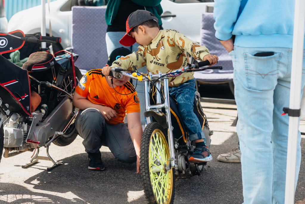Ett barn med keps och mönstrad skjorta sitter på en cykel med gula ekrar medan en vuxen sitter på huk i närheten och pekar på cykeln. Runt omkring dem syns andra människor och motorcyklar.