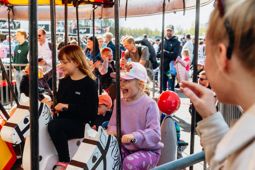 Två unga flickor ler och skrattar medan de rider på leksakshästar i en karusell på en utomhusmarknad, omgivna av andra barn och vuxna som njuter av evenemanget.