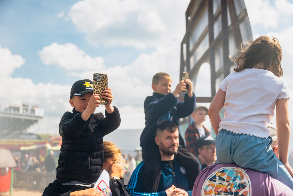 Barn som sitter på vuxnas axlar tar foton med smartphones vid ett utomhusevenemang. Ett barn bär keps och håller i ett leopardmönstrat mobilskal, medan en flicka i jeans vänder sig bort och klättrar på en lekställning. Ljus, solig himmel ovanför.