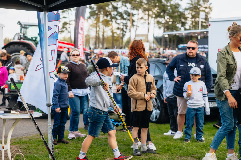 Barn och vuxna samlas utomhus vid ett välbesökt evenemang. Två pojkar går i förgrunden, den ena håller i en hockeyklubba. Människor står och pratar, med bilar och flaggor synliga i bakgrunden. Träd kantar området.