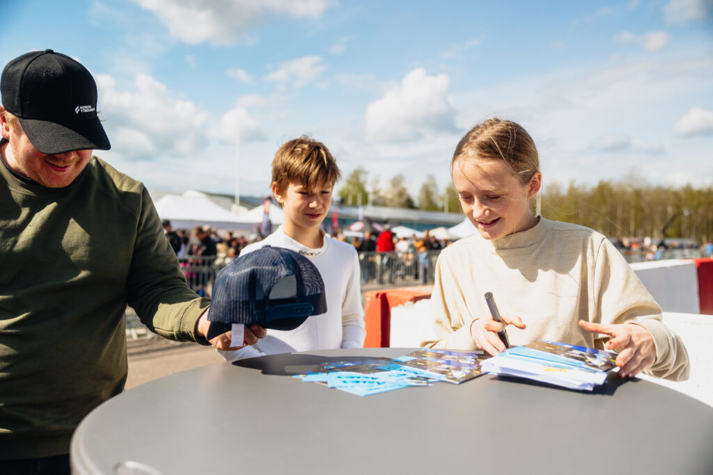 Två unga människor ler när den ena signerar foton vid ett utomhusevenemang. En man håller fram en keps för att få en autograf. Himlen är klar och det finns människor och tält i bakgrunden, vilket tyder på en livlig atmosfär.