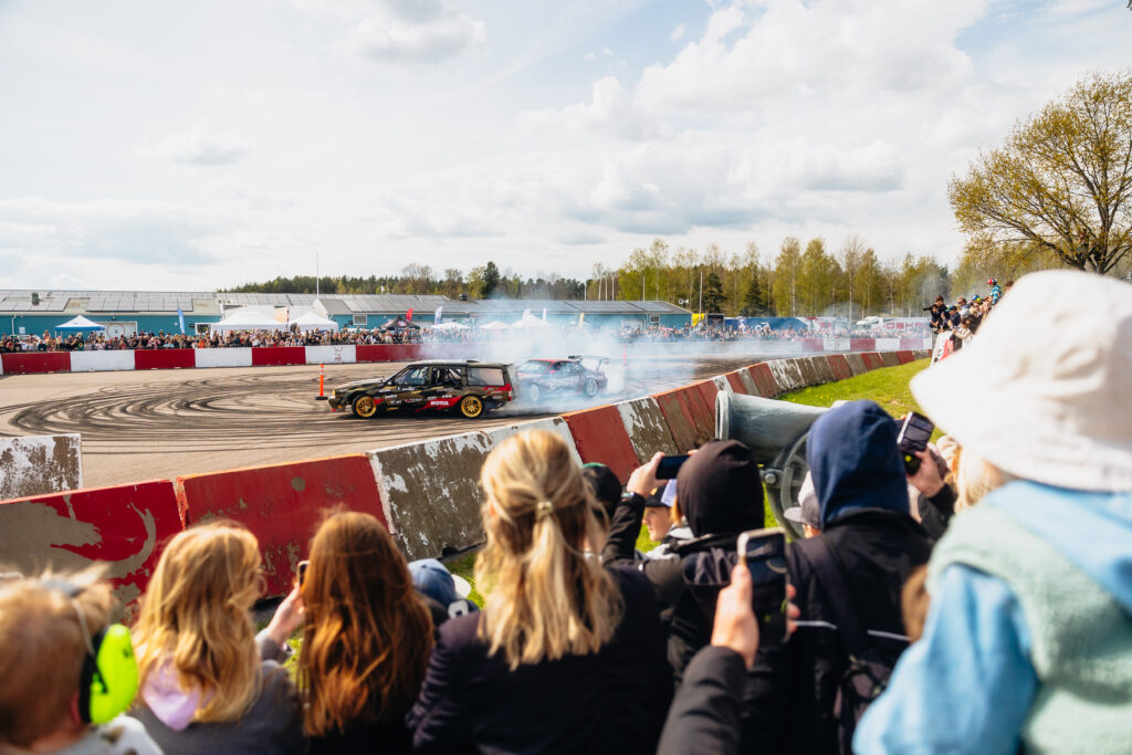 En folkmassa tittar på när två bilar driver runt på en racerbana och producerar rökmoln. Människor tar foton och videor, och däckmärken syns på asfalten. Evenemanget äger rum utomhus på en delvis molnig dag.