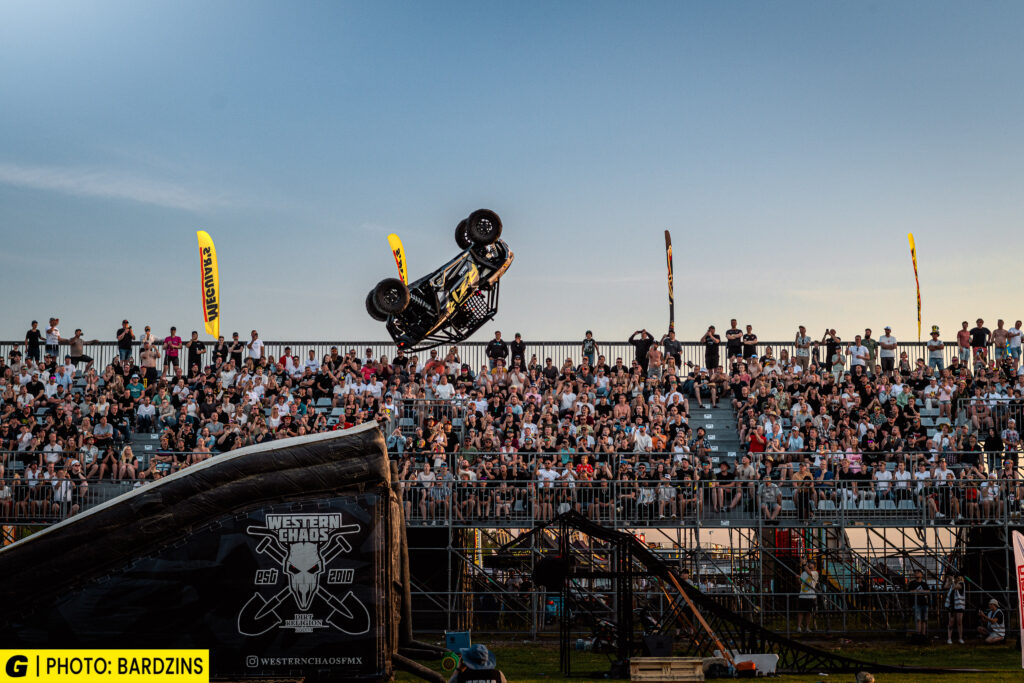 En stuntbil gör en voltning i luften ovanför en stor publik som sitter på läktare under ett utomhusevenemang i solnedgången. Evenemangsbanderoller och säkerhetsramper syns i bilden.
