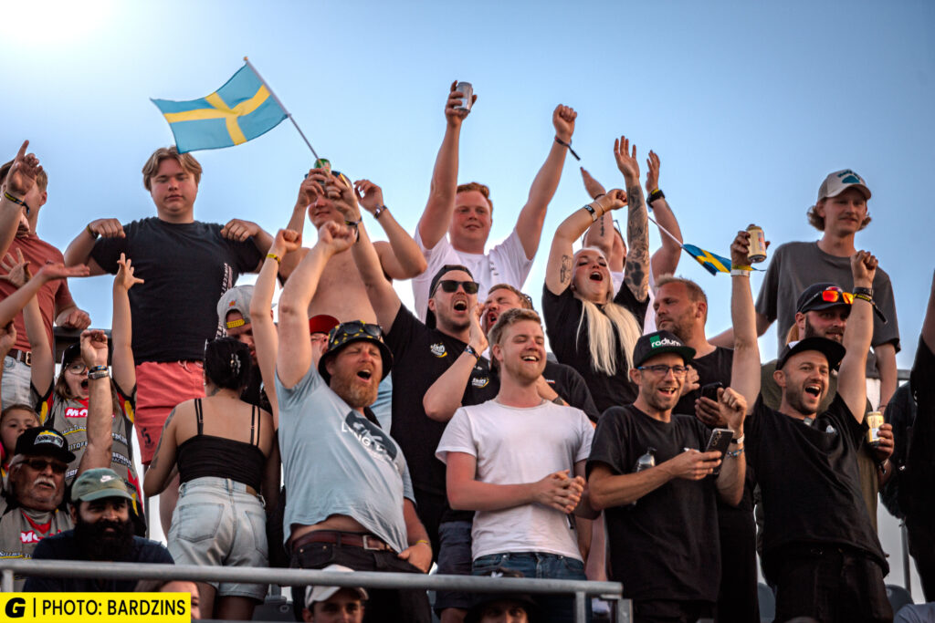 En jublande folkmassa, några med drinkar i handen och viftande med en svensk flagga, firar tillsammans utomhus under en klarblå himmel. Många har händerna uppsträckta i glädje.