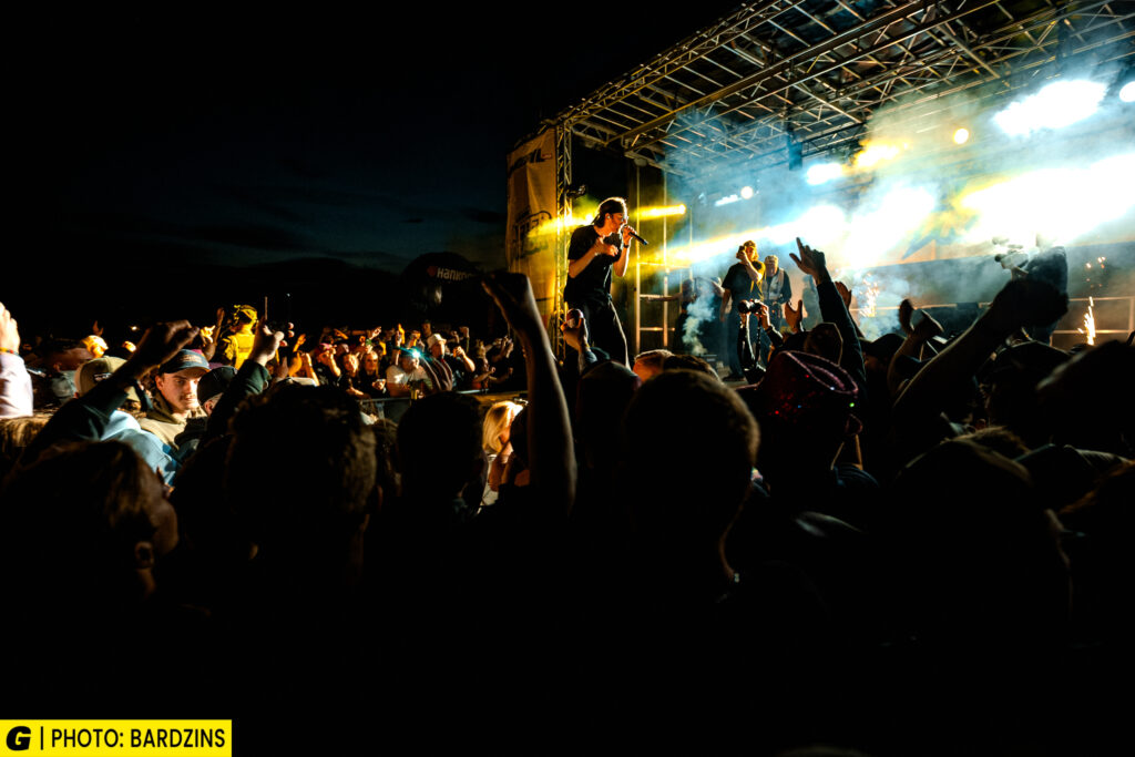En livlig konsertscen på kvällen med en publik som ser en sångare och ett band uppträda på en starkt upplyst utomhusscen. Några i publiken har händerna i luften och njuter av den energiska atmosfären.