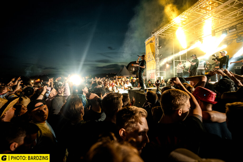 En stor publik njuter av en utomhuskonsert på kvällen, med starka scenljus som lyser upp artisterna på scenen och rökeffekter i luften. Människor i publiken har händerna i luften, vilket skapar en energisk atmosfär.
