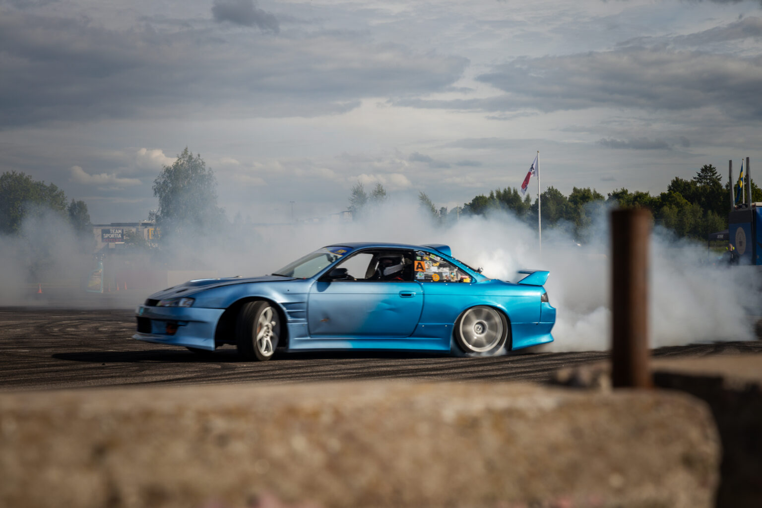 En blå sportbil driver på en asfaltsbana och skapar tjocka moln av vit rök under en delvis molnig himmel, med träd och flaggor synliga i bakgrunden.