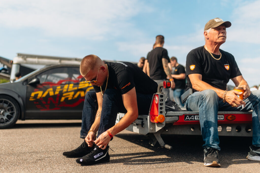 Två män sitter på ett bilsläp; den ene knyter sin sko medan den andre håller i en drink. Båda bär svarta skjortor och solglasögon. En modifierad racerbil med grafik syns i bakgrunden och flera personer står i närheten.
