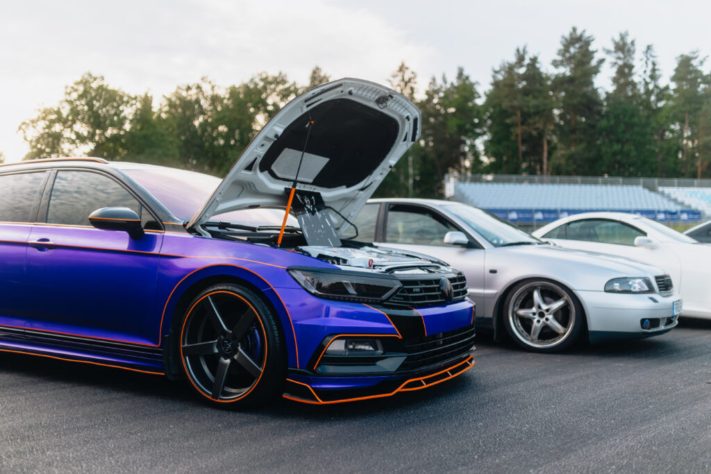 En blå och svart bil med orangefärgade detaljer har motorhuven öppen och står parkerad bredvid en vit bil på en utomhusparkering med träd och läktare i bakgrunden.