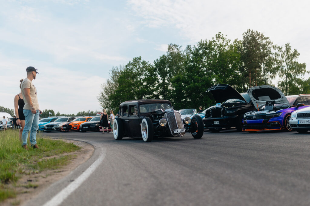 En svart veteranbil med vita hjul kör förbi en rad färgglada moderna bilar som står parkerade på en väg, vissa med öppna motorhuvar. Människor står i närheten och i bakgrunden syns träd under en delvis molnig himmel.