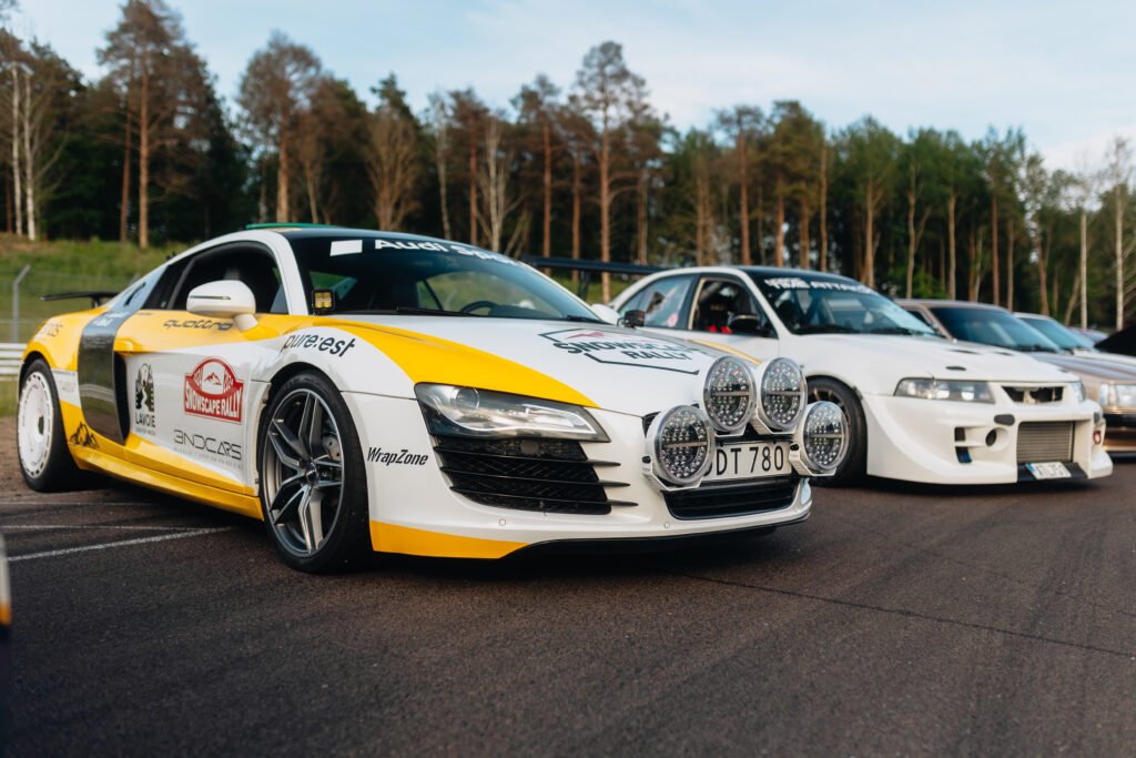 En gul och vit racerbil med stora rallylyktor framtill står parkerad på en väg bredvid en annan modifierad vit bil, omgiven av träd och grönska i bakgrunden.