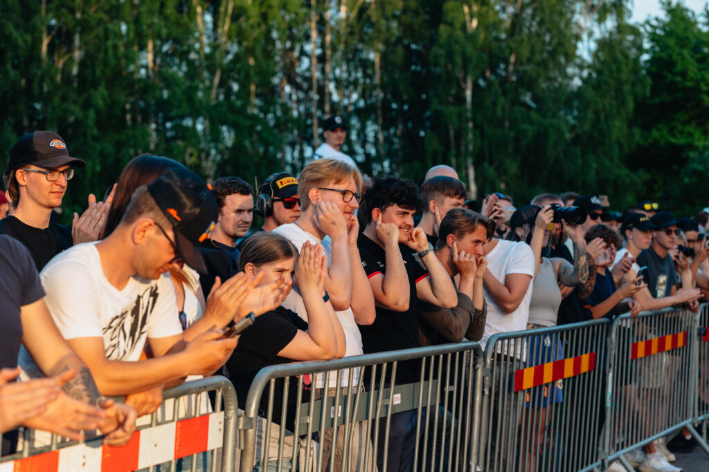 En grupp människor står tätt bakom en metallbarriär, vissa applåderar och andra håller för öronen, vid ett utomhusevenemang med träd i bakgrunden.