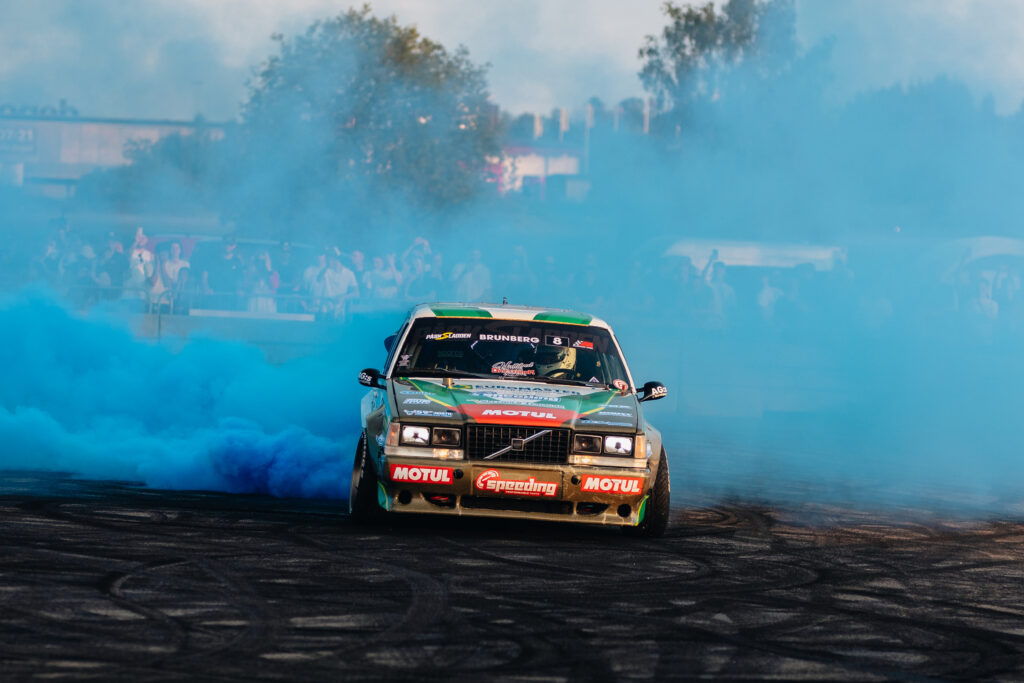 En grön racerbil driver runt ett hörn och skapar tjock blå rök från sina däck på en bana, med suddiga åskådare och träd som syns i bakgrunden.