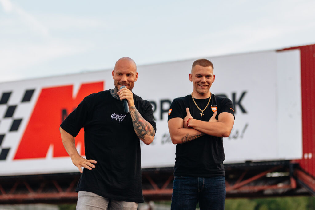 Två män står framför en stor skylt utomhus. Den ene, skallig och tatuerad, håller i en mikrofon och ler. Den andre står med armarna i kors och ler också. Båda bär vardagskläder och i bakgrunden syns en suddig reklamtavla.