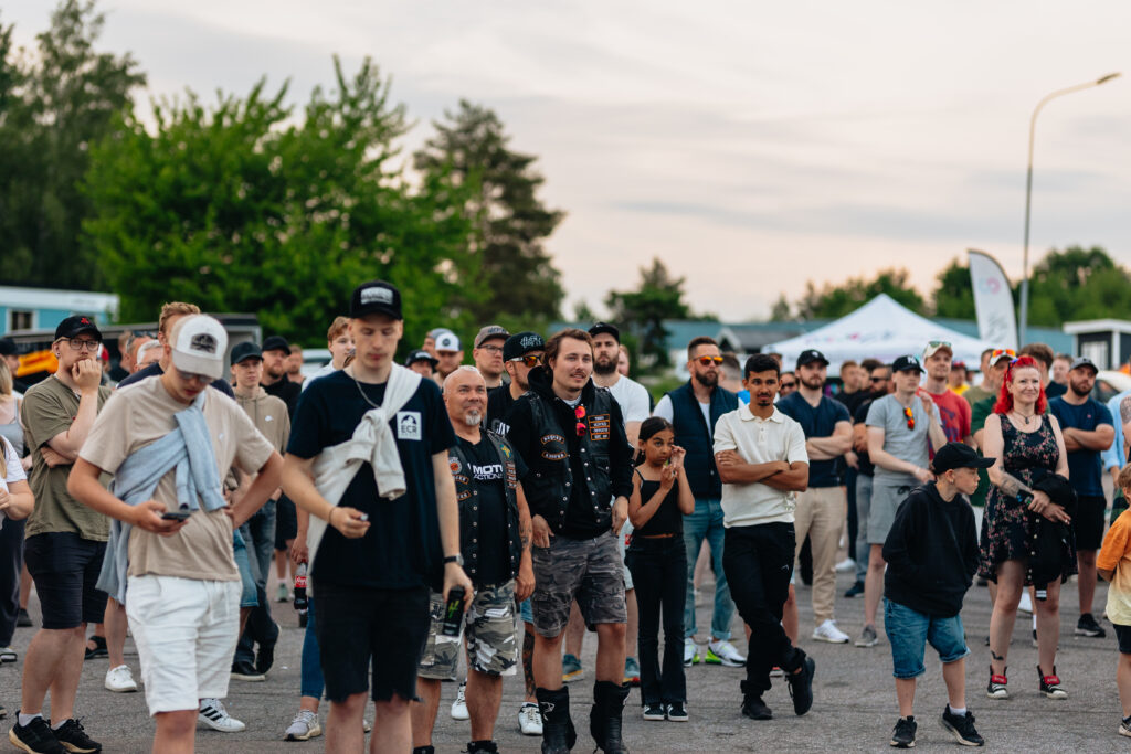 En grupp människor med olika bakgrund, män, kvinnor och barn, samlas utomhus i samband med ett evenemang. Några bär hattar och fritidskläder. I bakgrunden syns träd, tält och en molnig himmel i skymningen.