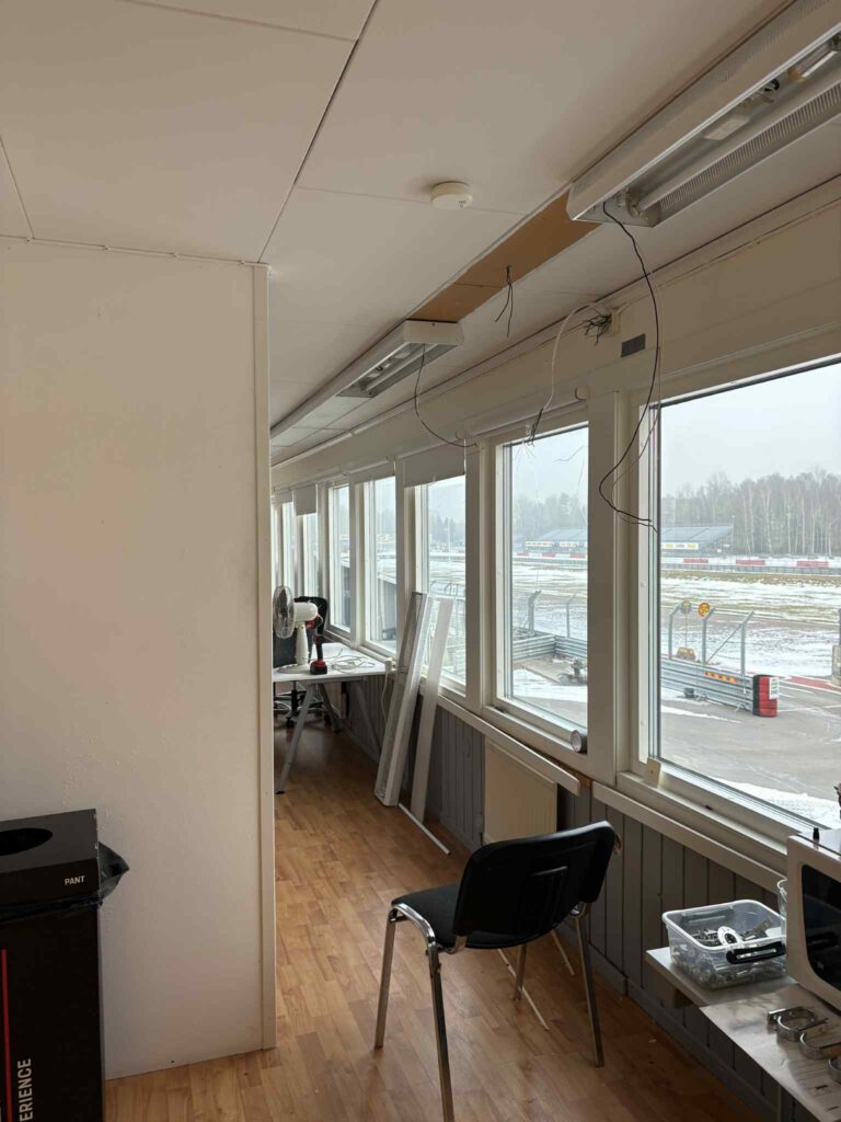 Ett rum med stora fönster som vetter mot en snöig utomhusmiljö, en svart stol, några utspridda föremål och nedhängande ledningar i taket. Golvet är av trä och rummet fylls av naturligt ljus.