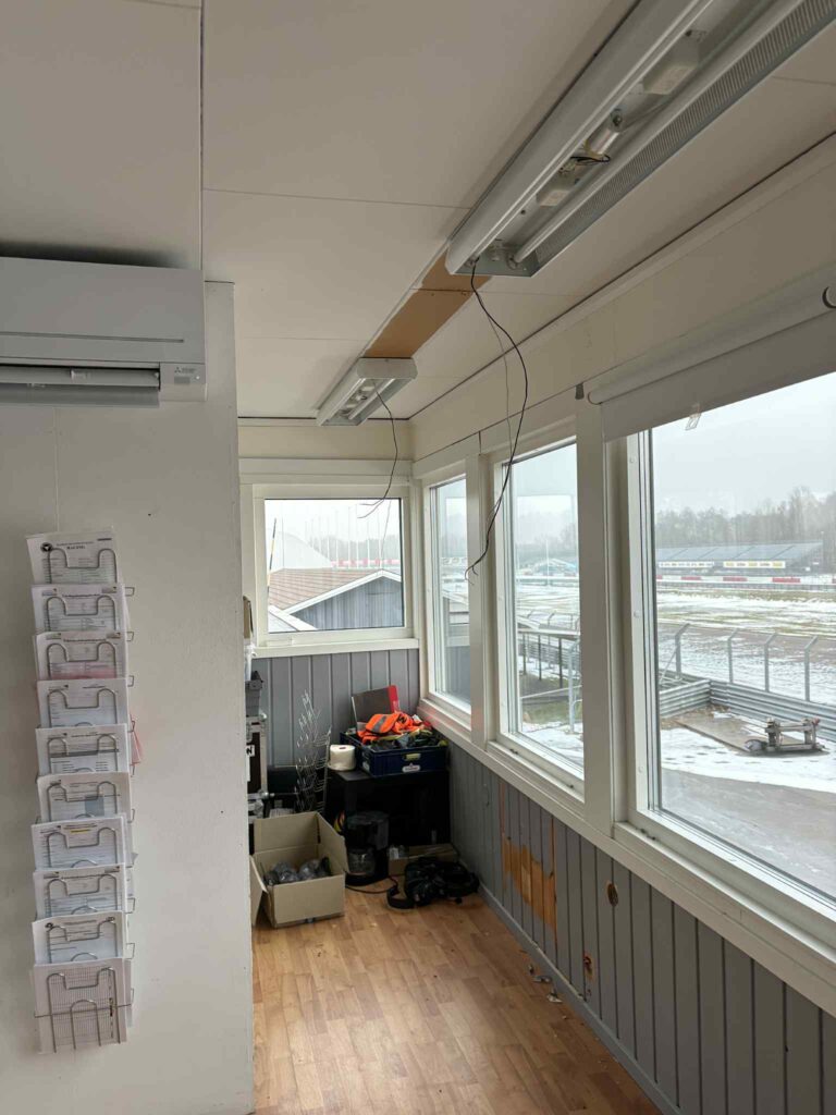 Ett litet kontorsutrymme med stora fönster, lysrör och trägolv. Verktyg och förnödenheter finns på ett skrivbord, en soptunna på golvet och en vägghylla med plastfickor till vänster. Utanför syns en snöig scen.