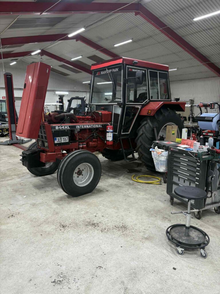 En röd International 844XL-traktor med öppen motorhuv står parkerad inne i en verkstad. Verktyg, utrustning och en rullande verktygslåda omger traktorn under ett högt, vitt och rött metalltak.