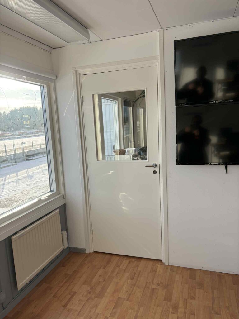 En vit dörr med ett fönster i ett ljust rum med trägolv, stora fönster till vänster, ett element under fönstret och två väggmonterade svarta skärmar till höger. Solljuset flödar in från utsidan.