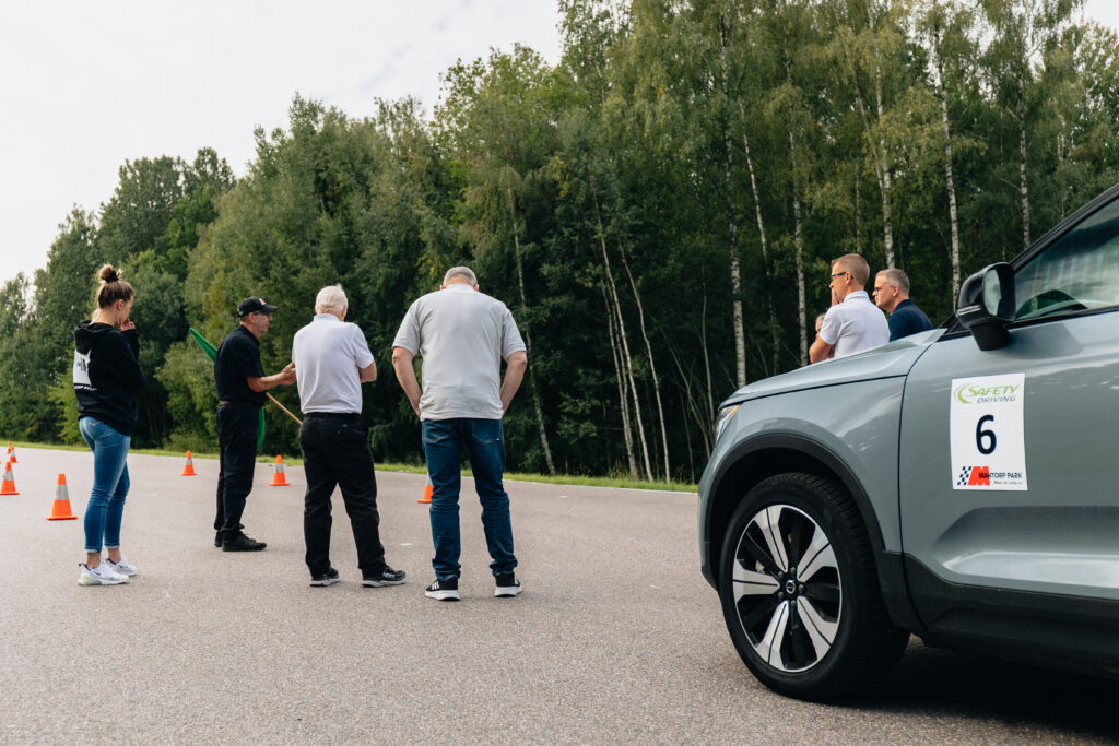 Flera personer står på en asfalterad väg nära orangea trafikkoner och en grå bil med siffran 6, omgiven av höga gröna träd, och verkar diskutera något utomhus.