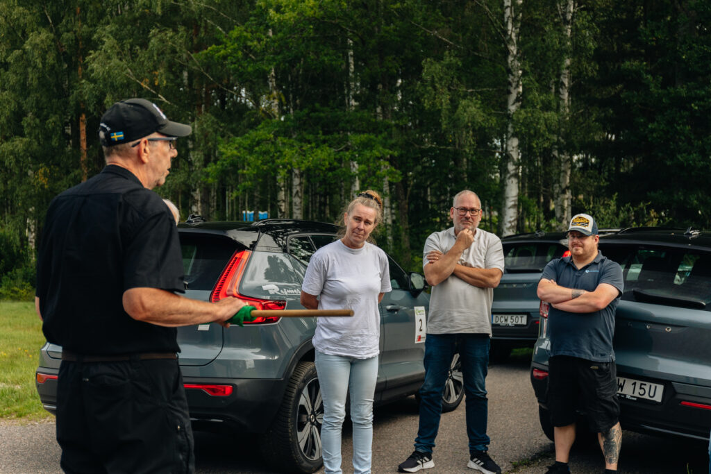 En svartklädd man håller i en käpp och talar till tre personer som står vid parkerade bilar på en väg. Gruppen lyssnar uppmärksamt, omgiven av grönska och träd i bakgrunden.