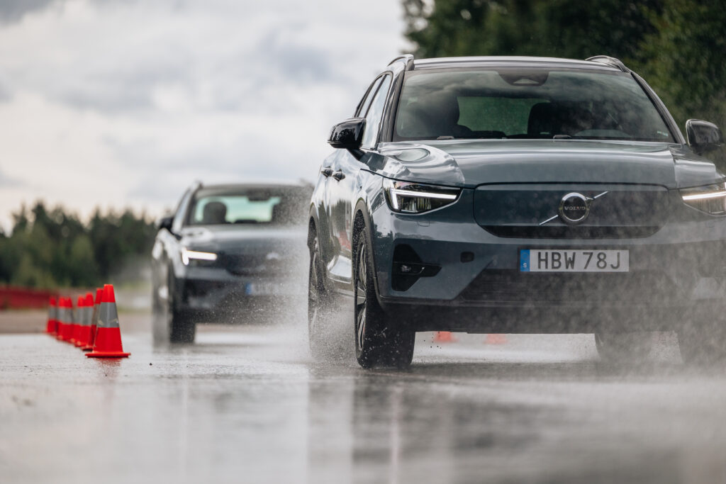 Två Volvo SUV:ar kör på en våt väg och stänker vatten medan de manövrerar runt orangea trafikkoner. Himlen är molnig och i bakgrunden syns träd.