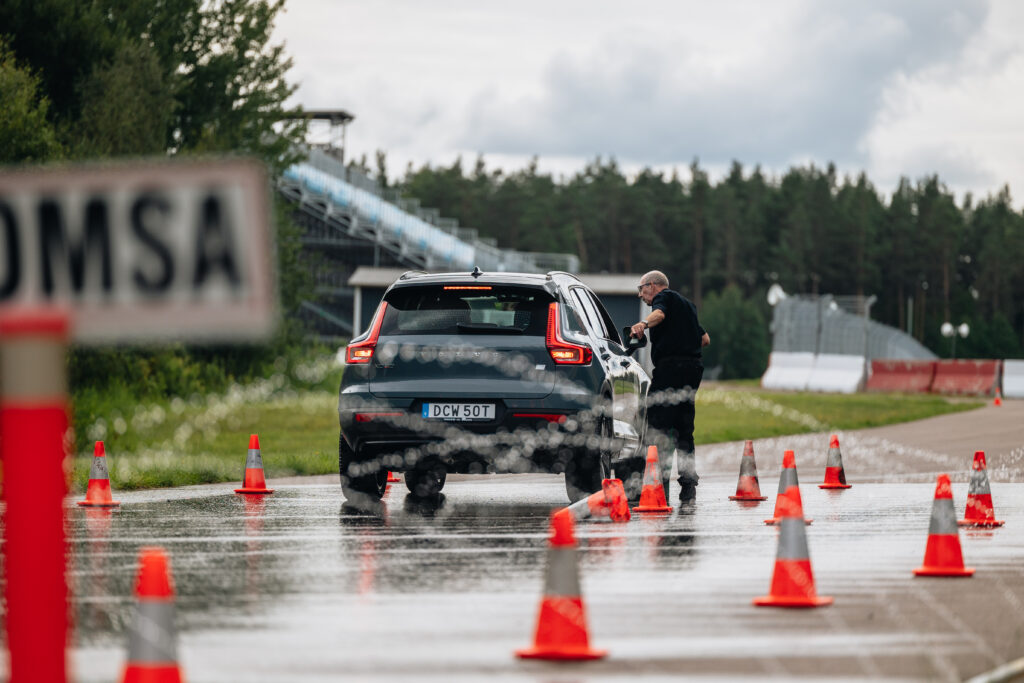 En person står vid förarsidan av en svart SUV på en våt, konbelagd bana, med en suddig OMSA-skylt i förgrunden och träd i bakgrunden.