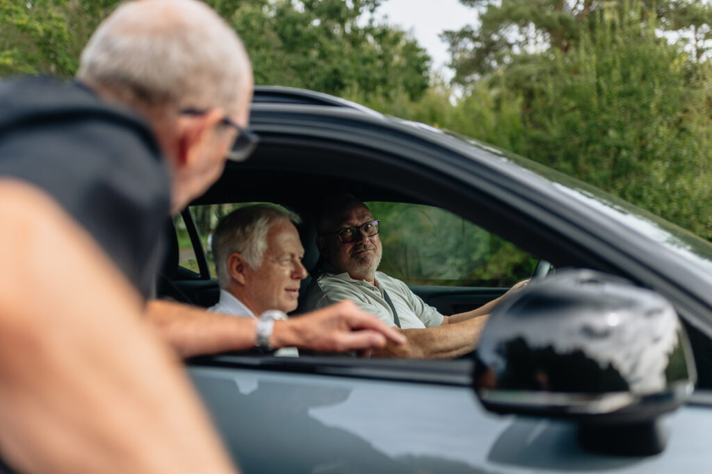 Två män sitter i framsätet på en bil medan en annan man står utanför, lutar sig in genom det öppna fönstret och pratar med dem. Bakgrunden är fylld av grönska och träd.