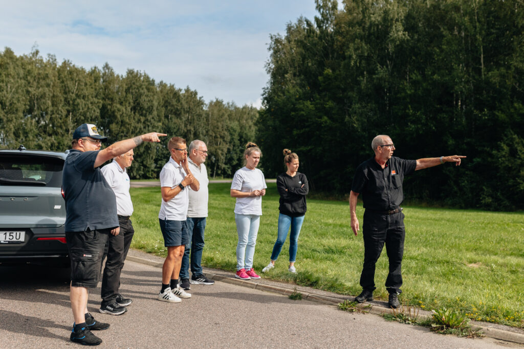 En grupp på sex personer står vid sidan av en väg nära en bil, och några pekar mot gräsytan och träden i fjärran under en ljus himmel.