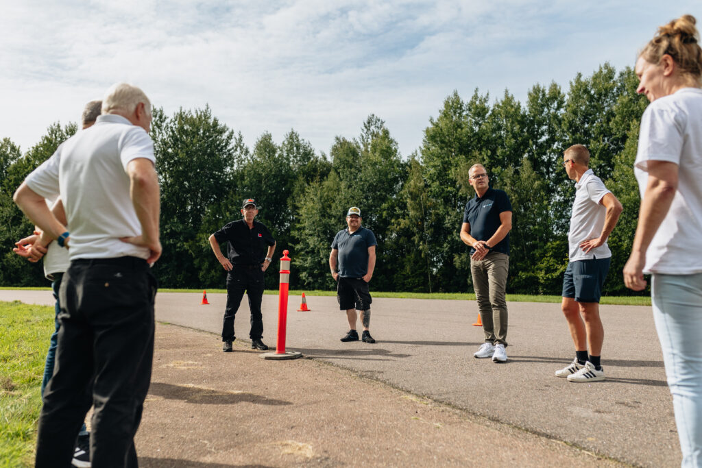 En grupp människor står i en cirkel på en asfalterad utomhusyta med träd i bakgrunden och lyssnar på en svartklädd man som står nära en orange trafikkon en solig dag.