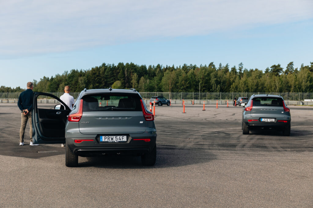 Två grå Volvo SUV:ar står parkerade på en asfalterad parkeringsplats nära orangefärgade trafikkoner. Två män står vid den ena bilen, varav den ena håller dörren öppen. Träd och ett staket syns i bakgrunden under en blå himmel.