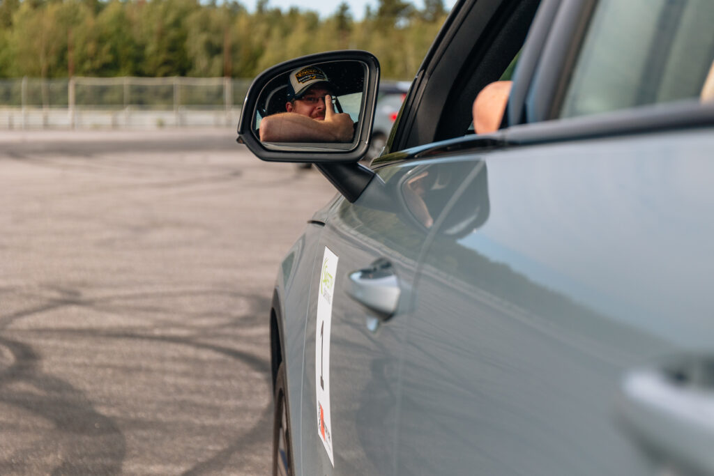 En man med keps speglas i sidospegeln på en parkerad grå bil på en asfalterad tomt, med träd och ett staket i bakgrunden.