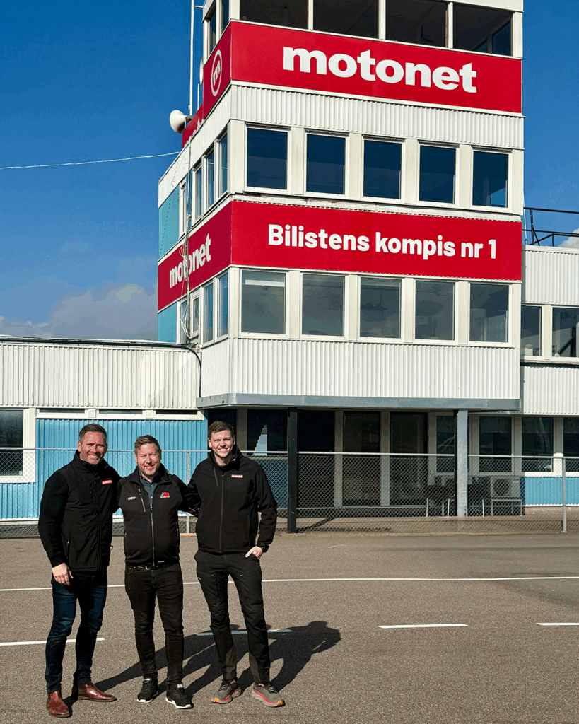 Tre personer står leende framför en motorsportbyggnad med stora röda motonettskyltar och texten Bilsitens kompis nr 1. Vädret är soligt och gruppen är klädd i svarta kavajer.