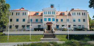 En stor vit byggnad med rött tak, som kallas Stadshotell ovanför entrén, har symmetriska fönster och en central trappa som leder upp till ytterdörren. Uteserveringar är synliga på båda sidor.