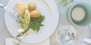 En vit tallrik med pocherad lax, färskpotatis, sparris, gröna bönor, en citronklyfta och en dillkvist. På tallriken vilar en gaffel och en kniv. Ett stearinljus och ett vinglas står på bordet.