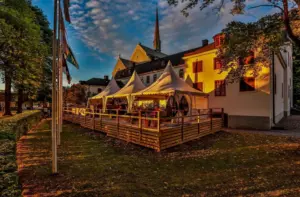 En mysig uteservering med vita tält ligger intill en historisk byggnad i solnedgången. Byggnaden är upplyst av ett varmt ljus och i bakgrunden skymtar en kyrktornsspira under en delvis molnig himmel.