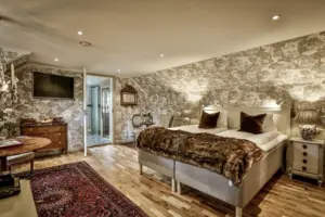 Ett mysigt sovrum med mönstrad tapet, en stor säng med pälsmössa, två sängbord med lampor, en rödmönstrad matta på trägolv och en byrå. En öppen dörr avslöjar ett ljust badrum.