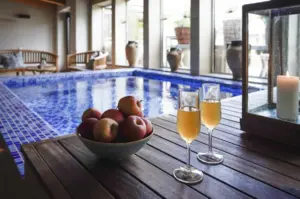 En skål med äpplen och två glas mousserande dryck står på ett träbord vid en inomhuspool, med ett tänt ljus i en lykta i närheten och naturligt ljus som flödar in genom stora fönster.