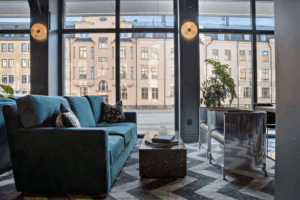 Modernt vardagsrum med en tealfärgad soffa, soffbord i marmor, växter och stora fönster som visar klassiska beige byggnader utanför. Solljuset flödar in och skapar en ljus och inbjudande atmosfär.