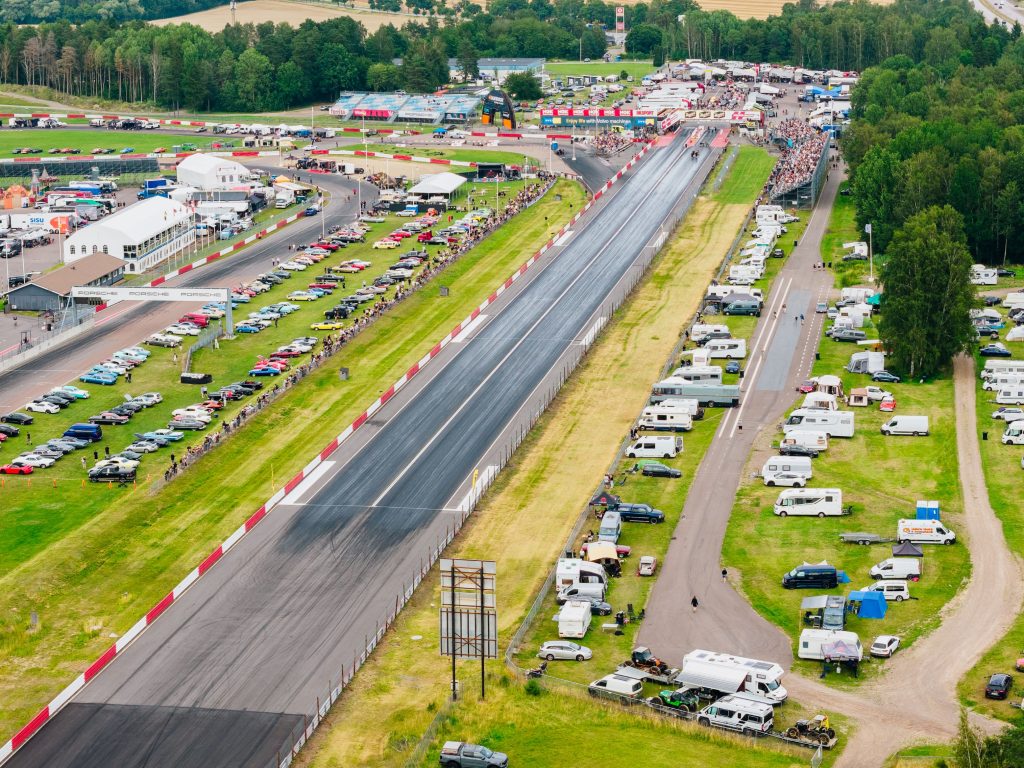 Flygbild över en dragracingbana med uppställda bilar, åskådare på läktarna, husbilar och tält uppställda längs sidorna samt gröna träd som omger området.